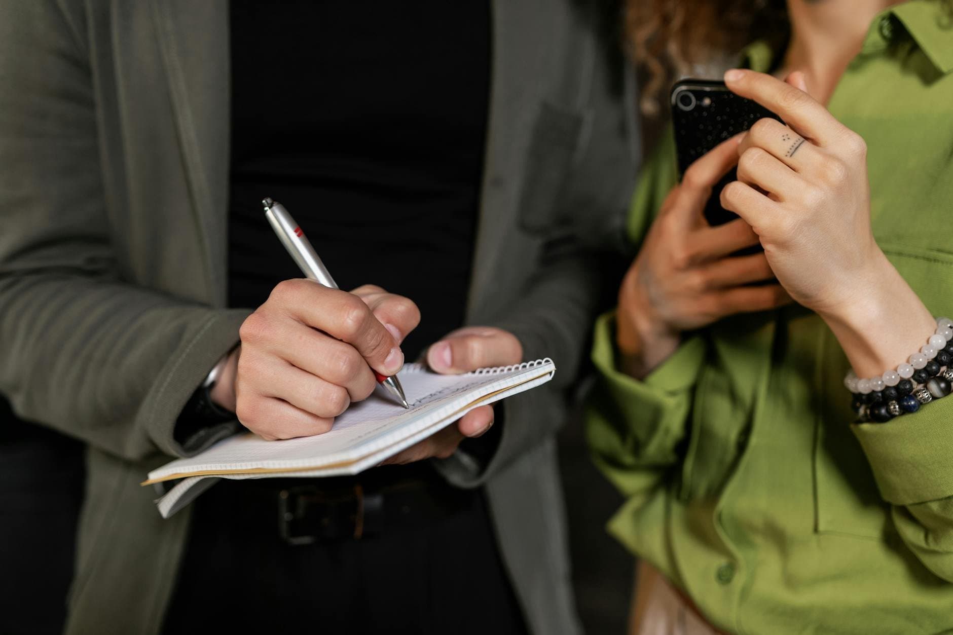 close up of person writing in a journal with a smartphone nearby (Pexels)